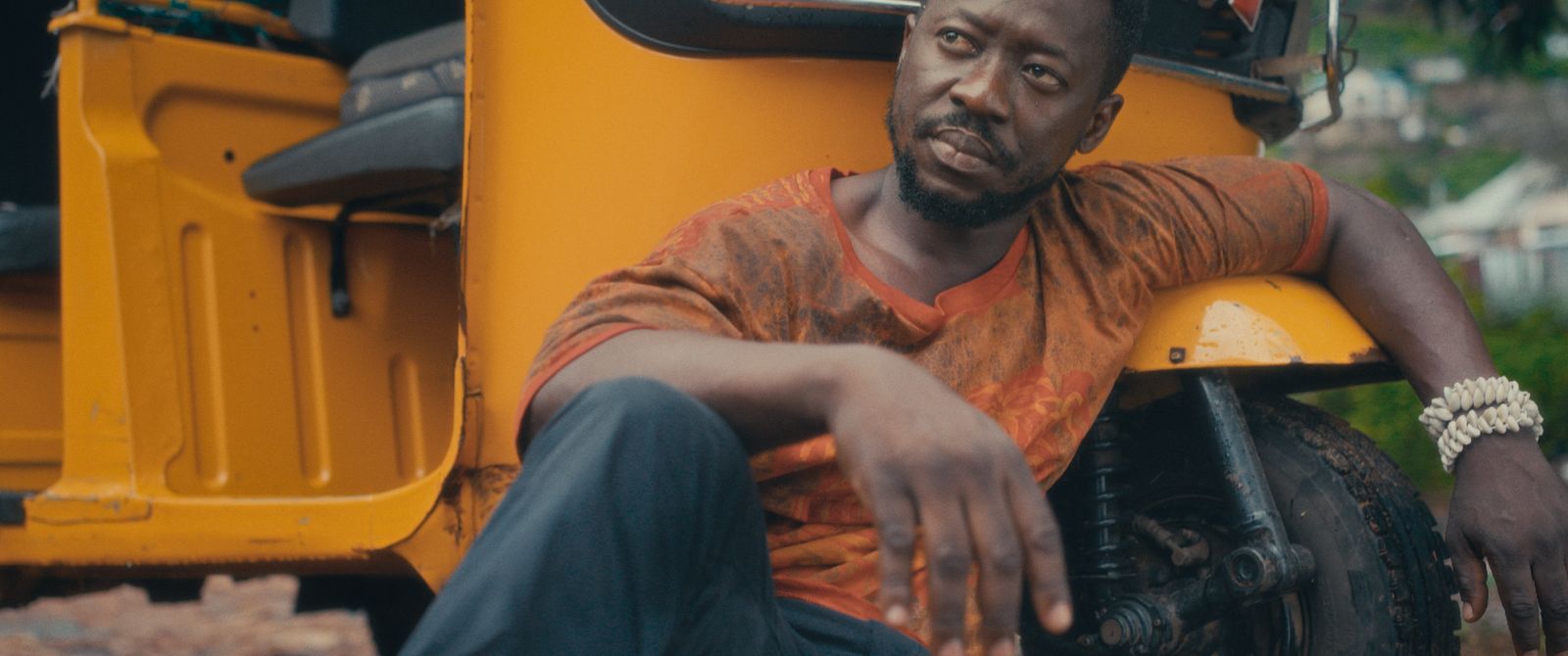 A still from ARRIES: a man seated against a yellow three-wheeled taxi, a beaded band at his wrist, hills behind him.